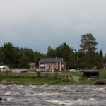 Röd byggnad vid forsande flod, omgiven av skog, fordon och flagga under mulen himmel.