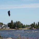 Person kitesurfar på sjö med hus och träd i bakgrunden under blå himmel.