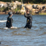 Två personer i våtdräkter och hjälmar i vatten, en med kite-lina, strand och stenar synliga.