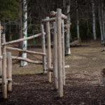 Utomhuslekplats med träklätterredskap omgiven av björkar i skogsmiljö med mark av bark.