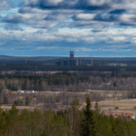 Landskap med skog, fält, byggnader och en industriell anläggning under en molnig himmel.