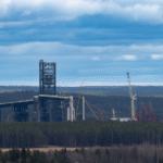 Industriell anläggning under konstruktion med flera kranar vid skogskant och molnig himmel.