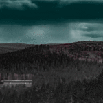 Vy över skog med klippa och långt byggnad under mörk och molnig himmel.