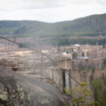 Klippkant med rostiga metallstavar och utsikt mot skog och by i bakgrunden.